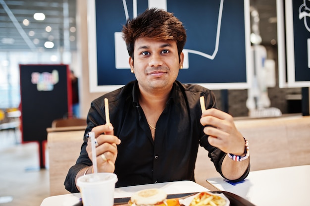 Stylish indian man sitting at fast food cafe and eating french fries.