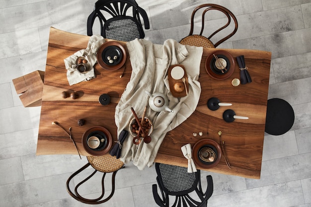 Stylish interior design of dining room with wooden walnut table, retro chairs, tableware, plates, tablecloth, teapot, food, decoration and elegant accessories. Cement floor. Top view.