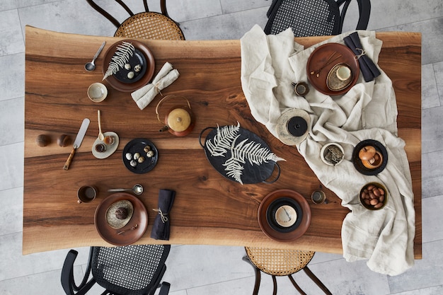 Stylish interior design of dining room with wooden walnut\
table, retro chairs, tableware, plates, tablecloth, teapot, food,\
decoration and elegant accessories. cement floor. top view.