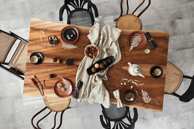 Stylish interior design of dining room with wooden walnut table, retro chairs, tableware, plates, tablecloth, teapot, food, decoration and elegant accessories. Cement floor. Top view.