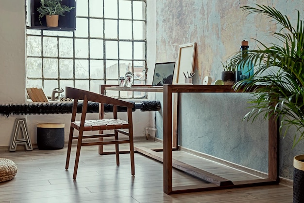 나무 책상, 의자, 사무용품, 노트북, 식물, 램프 및 우아한 액세서리를 갖춘 로프트 아파트의 세련된 인테리어 디자인. 현대 홈 오피스 장식입니다. 밝은 공간. 주형.