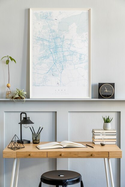 Stylish interior of home office room with black mock up poster map, wooden desk, black stool, clock, books, plants, cacti, office supplies, lamp and personal accessories in modern home decor.