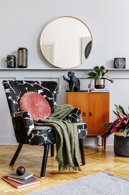 Stylish interior of living room with design armchair, wooden vintage commode, round mirror, shelf, plants, book, decoration, lantern, grey wall and persnoal accessories in home decor.