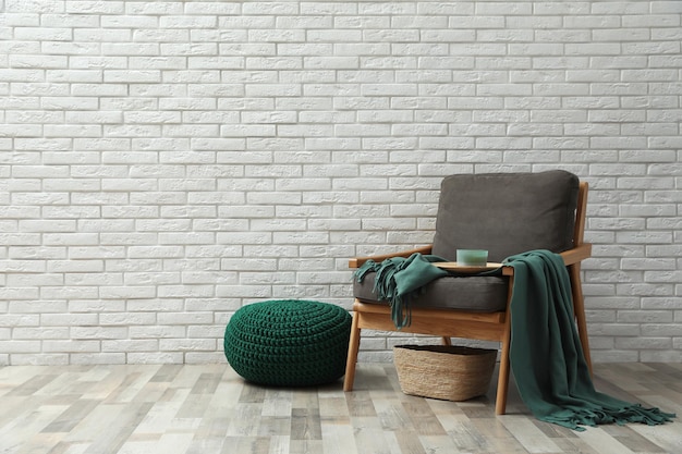 Stylish knitted pouf and comfortable armchair near white brick wall indoors space for text Interior design