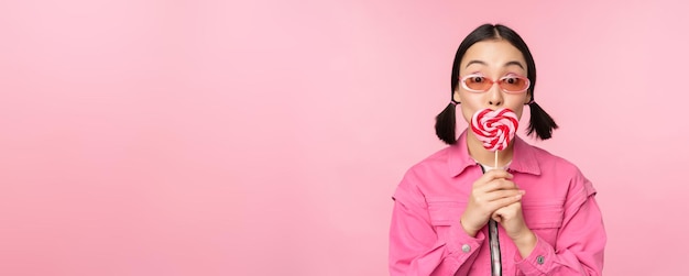 Stylish korean girl licking lolipop eating candy and smiling standing in sunglasses against pink background