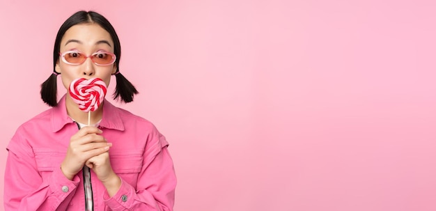 Stylish korean girl licking lolipop eating candy and smiling standing in sunglasses against pink background