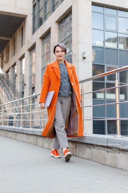 Stylish lady in bright orange coat in a city scene, using a laptop computer and working on it during a break. Outdoors technology. drinking coffee, tea, cocoa