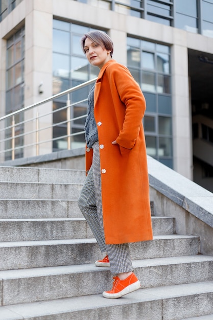 Stylish lady in bright orange coat posing on stairs