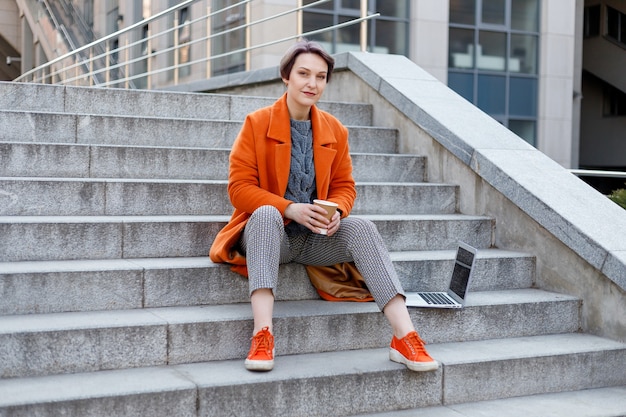 Stylish lady in bright orange coat with her laptop and coffee cup