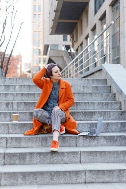 Stylish lady in bright orange coat with her laptop and coffee cup