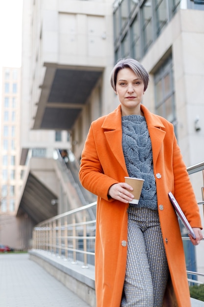 Stylish lady in bright orange coat with laptop and coffee cup