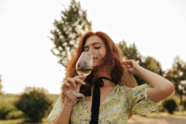 Stylish lady with red long hair and black bandage on her neck in green dress and straw hat drinking champagne and posing outdoor