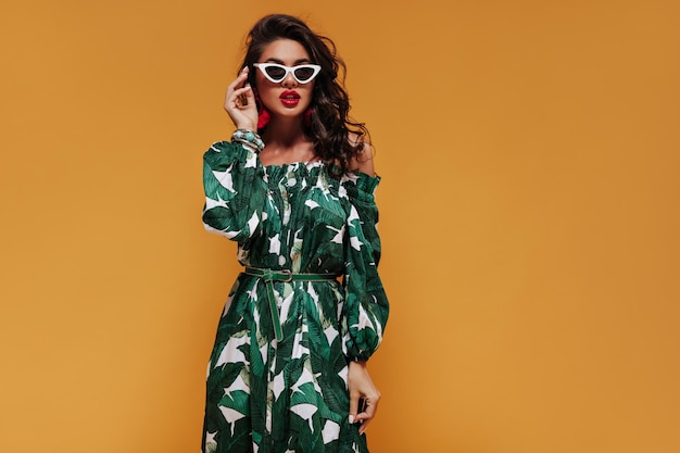 Stylish lady with wavy hair and white sunglasses in green dress with belt looking into camera on isolated orange backdrop