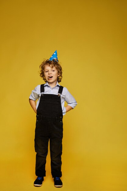 Stylish little boy in overalls and birthday cap posing at the yellow background, isolated. Kids fashion