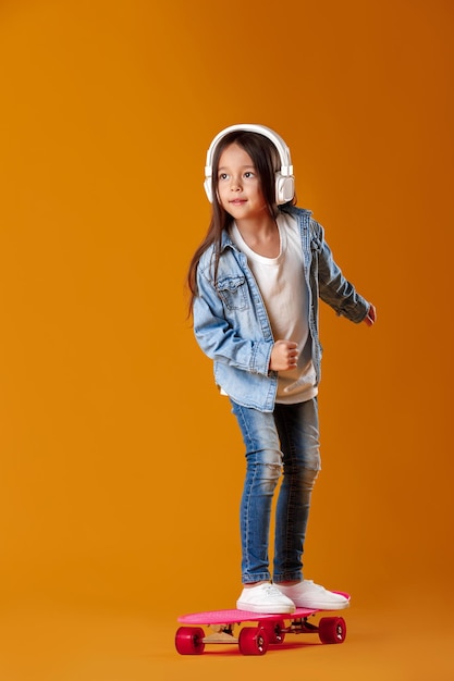 Stylish little child girl in headphones with skateboard in jeans clothes on orange background