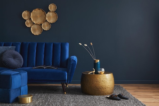 Stylish living room interior composition with velvet blue sofa\
golden side table pouf pillows and elegant home decor dark blue\
wallpaper template copy space