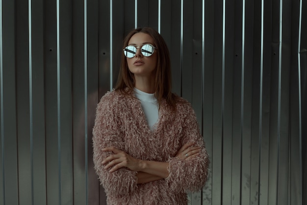 Stylish look up fashion tall model long brown hair woman in sunglasses with mirror effect in pink coat and white shirt poses laying with hands crossed on industrial storage parking metallic background