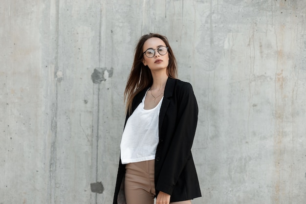 Stylish lovely young woman in fashionable beautiful outfit in trendy glasses poses near vintage building outdoors. Summer casual elegant clothes for women.