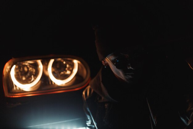 Stylish man in a black leather jacket with fashionable sunglasses sits near a headlight in the dark