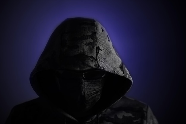 Uomo alla moda con cappuccio scuro occhiali neri e maschera sfondo scuro in luce blu hacker attaccante e bandito