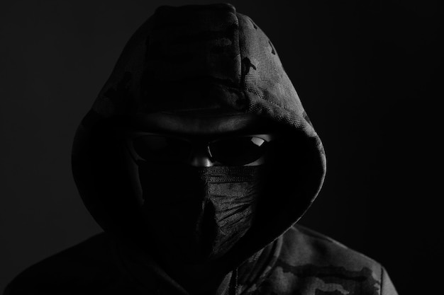 Stylish man in dark hoodie with black glasses and mask dark background hacker attacker and bandit