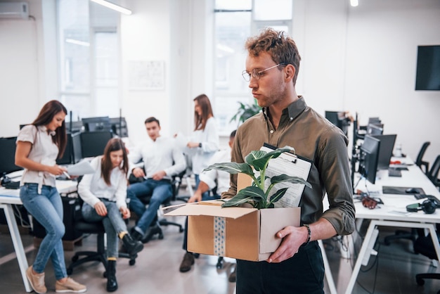 Stylish man in glasses holding box with plant in front of his colleagues in the office.