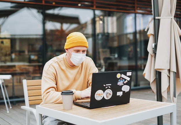 Stylish man hipster in face mask freelancer working on laptop in the street cafe new normal