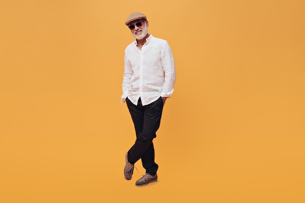 Stylish man in sunglasses cap and white shirt poses on orange background Fashionable adult with beard posing on camera with his hands in his pocket