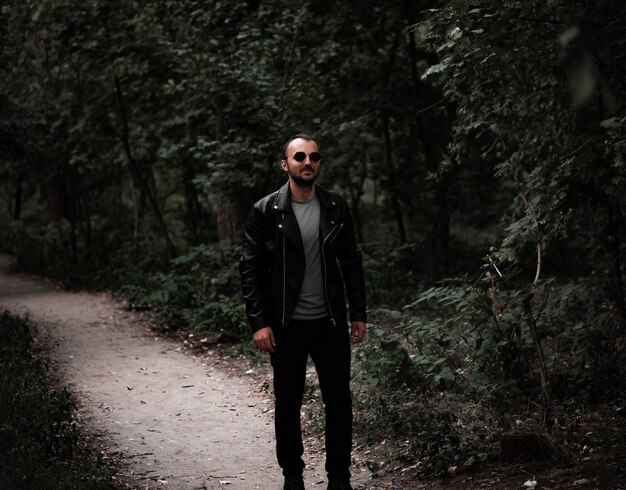 Stylish man in sunglasses and a leather jacket stands against the background of the forest