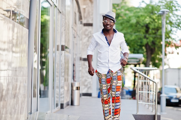 Stylish man in white shirt and colored pants with hat and glasses posed outdoor