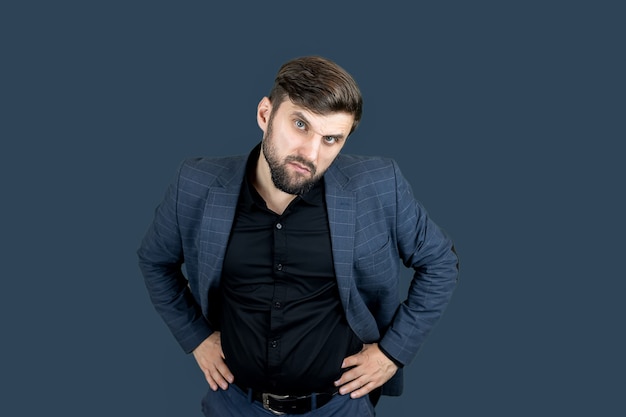 A stylish man with a beard in a blue suit holds his hands on his belt and looks at the camera