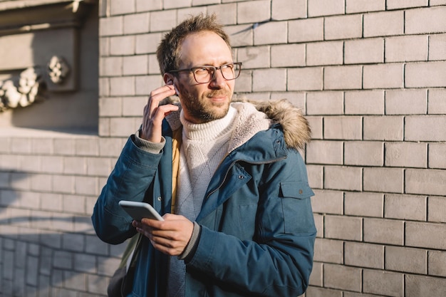 Stylish man with beard and glasses dressed in blue jacket inserts wireless headphones into ears