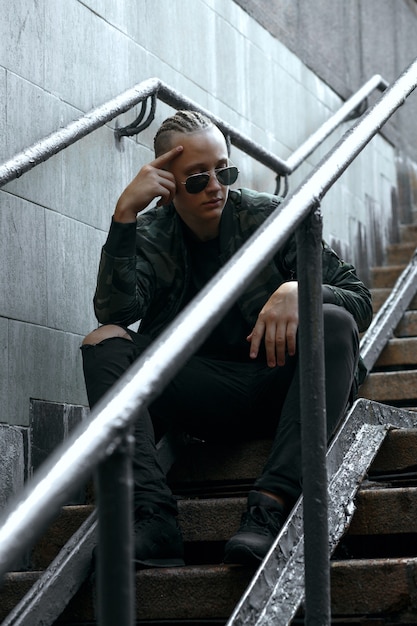 Stylish man with braids wears sunglasses and khaki jacket sitting on the street stairs