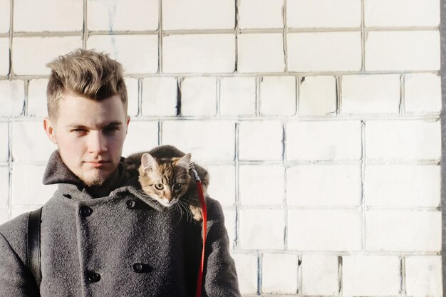 Stylish man with cute kitty on shoulder posing on background f brick wall in sunny street funny emotions space for text