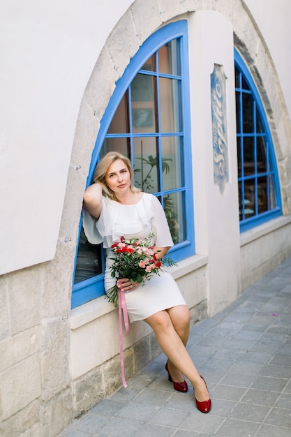 Stylish mature lady in white elegant dress posing at camera with flower bouquet outdoors in the city. Blue vintage window in ancient city on background.