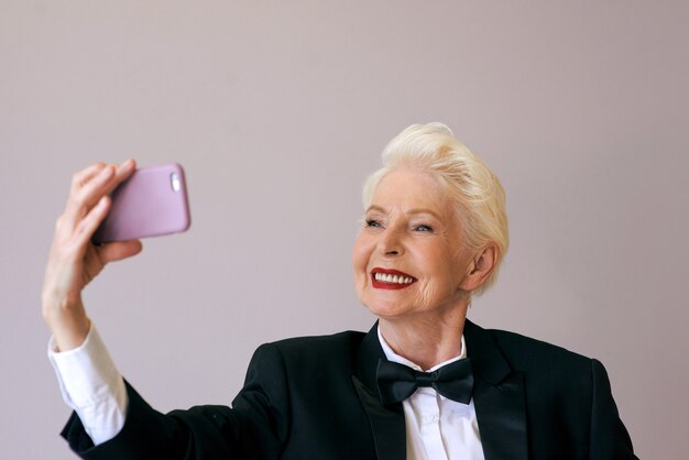 стильная зрелая старшая женщина в смокинге с мобильным телефоном, видеозвонком или селфи