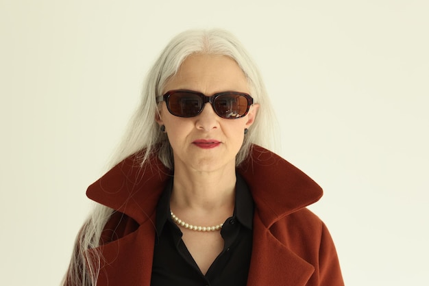 Stylish mature woman in sunglasses looks in camera on beige background portrait of greyhaired