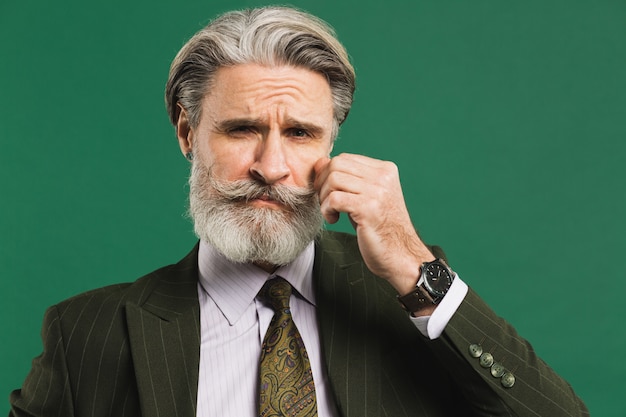 Stylish middle-aged man in suit fixes mustache and beard on green wall