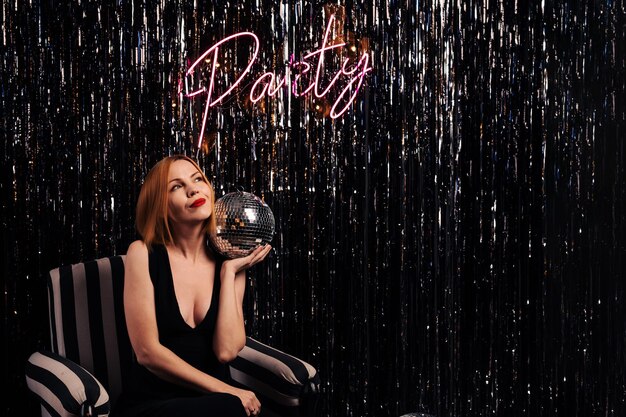 Stylish middle aged woman smiling and holding a mirror ball a\
woman of forty years old clubbing nightlife celebration\
entertainment concept party inscription on the luminous wall woman\
and party