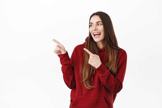 Stylish millennial woman pointing and looking aside at left copy space, smiling pleased, showing way to cool promo deal, special discount sale in store advertisement, standing over white wall