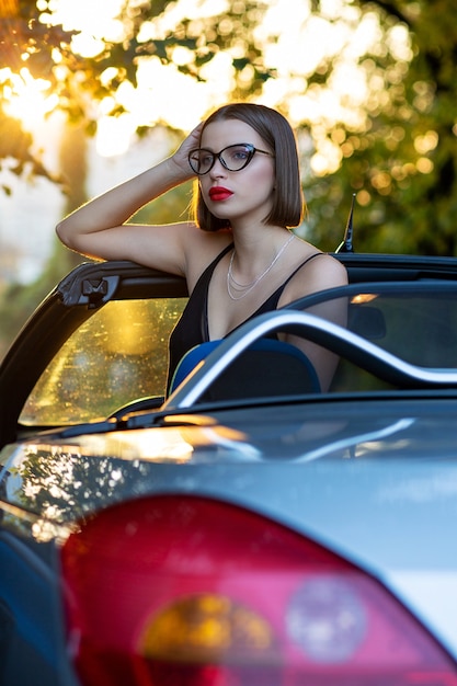 Stylish model wearing glasses, sitting in a convertible car with gold evening sunlight