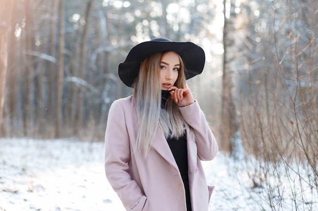 Stylish modern beautiful young woman in a knitted vintage dress in a stylish black hat in a pink elegant coat posing in the woods