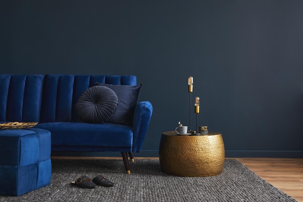 Stylish modern living room interior design with glamour blue\
velvet sofa pouf golden metal side table and modern home\
accessories dark blue wall template copy space