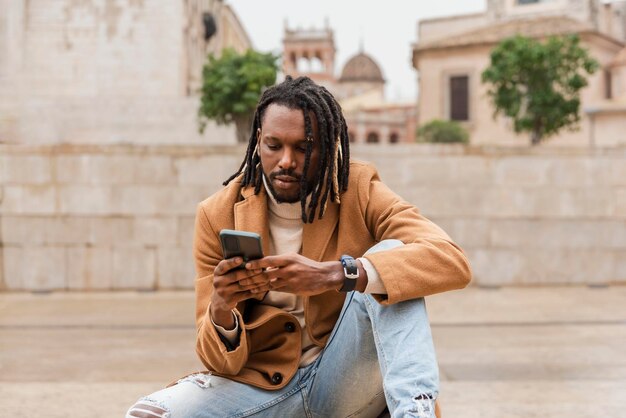 A stylish modern man with dreadlocks sitting in the city outdoors using smartphone