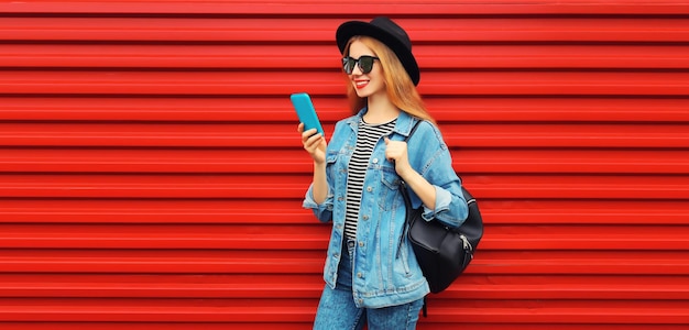 Stylish modern young woman with smartphone in jean jacket black round hat on red background