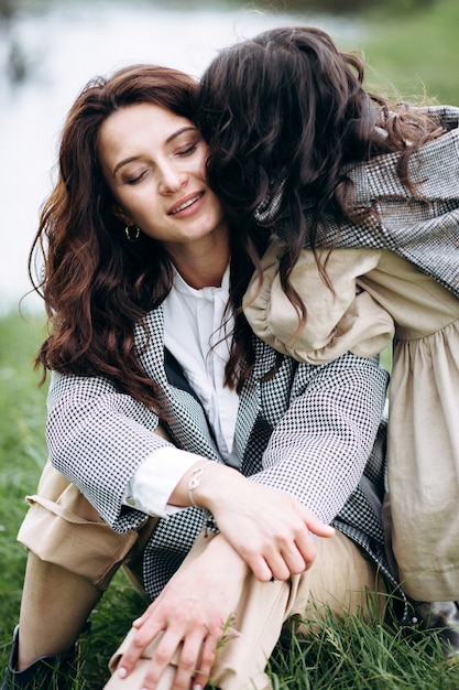 Stylish mother and daughter have fun outdoors in a field with green grass by the river when it rains