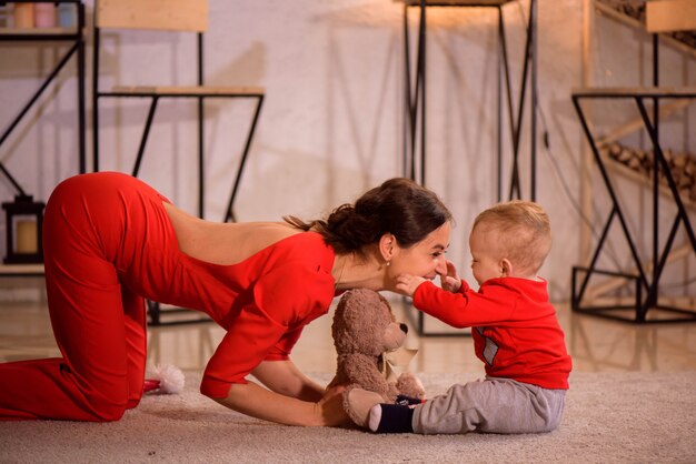 Stylish mother playing with baby boy in room. Sitting on floor near Christmas decorations.