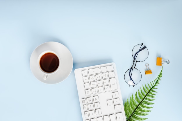 Stylish office desktop with keyboard cup of tea glasses and a plant on a blue background top view