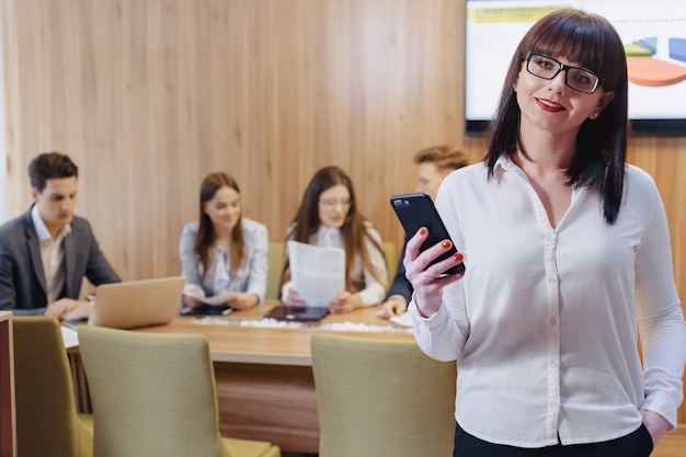 Stylish office worker woman in glasses with phone in hands against background of working colleagues