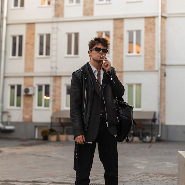 Stylish portrait of a young man with cigarette in fashion sunglasses in cool leather black jacket with a trendy backpack on a city street. Urban hipster guy in wear in american style smokes outdoors